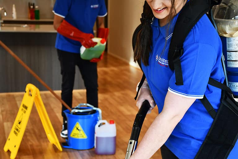 A cleaner selecting the right cleaning product for floor mopping, while the other takes care of vacuuming the floors