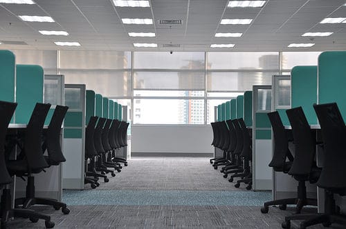 Office desks with black chairs and partitions, bright natural light through large windows.