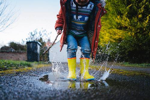 Child wearing rain boots playing in puddles outdoors.